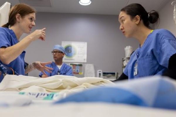 Two women lean across a hospital bed. The woman on the left gestures at the woman on the right. A man in the background looks on.