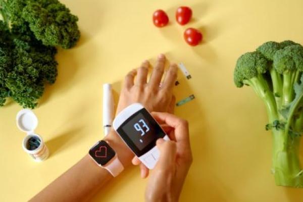 A person’s hand is surrounded by three types of healthy food, kale to the left, tomatoes above and broccoli to the right. Also shown are diabetes-testing strips, a container of strips and a glucose-testing device and electronic monitor, which displays a blood-sugar level of 93.