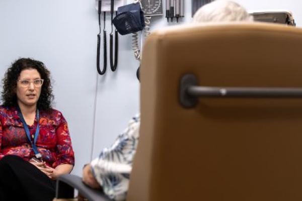 Dr. Sarah Kawasaki talks to a patient seated in an office exam chair. The patient is shown from behind. A blood pressure cuff and other medical instruments hang on the wall nearby.