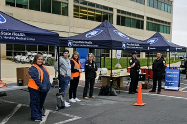 Three canopies branded Penn State Health Milton S. Hershey Medical Center a set up along the road in front of the medical center. Three hospital staff and three law enforcement officers stand under and around the canopies, facing the road.