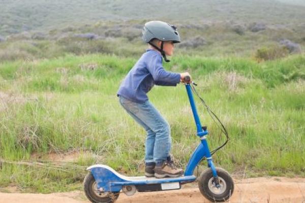 A young boy wearing a helmet rides an electric scooter on a dirt path.