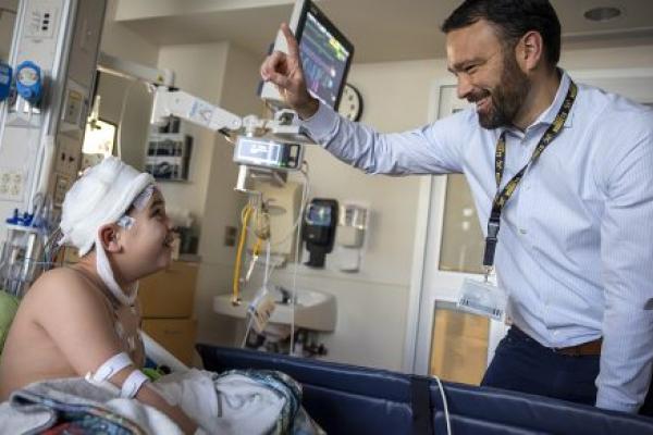 Dr. Brent O'Neill, right, wearing a shirt and tie, raises a finger and smiles at a boy in a hospital bed. The boy is looking up at him. The boy’s head is covered in bandages, he has an IV in his arm and has a blanket on him. Behind them are a monitor and medical equipment.