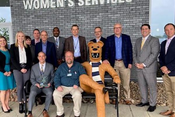A man in a suit sits on a bench outside Penn State Health Hampden Medical Center holding a glass award. Another man in street clothes sits beside him. A permanently affixed life-size statue of the Penn State Nittany Lion is also seated on the bench. A large group of men and women including Penn State Health and FM Global leaders stand behind the bench.
