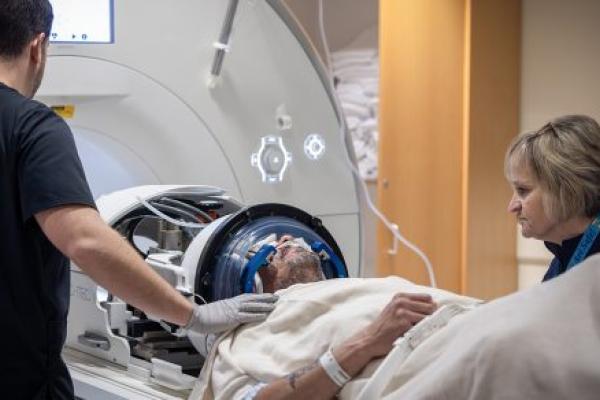 Two MRI technologists stand next to a male patient who is about to go into an MRI machine. The patient has a headgear on his head and a nasal cannula under his nose.