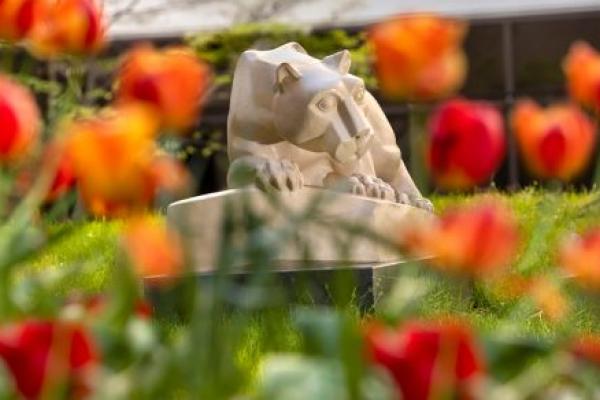 A statue of the Penn State Nittany Lion among grass and flowers and a building in the background.