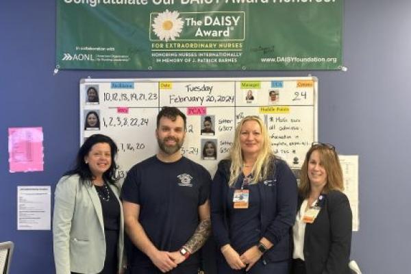 Four people stand side-by-side, smiling against a wall with a marker board, and a sign that says, “Congratulate Our DAISY Award Honoree! The DAISY Award for extraordinary nurses. Honoring nurses internationally in memory of J. Patrick Barns.”