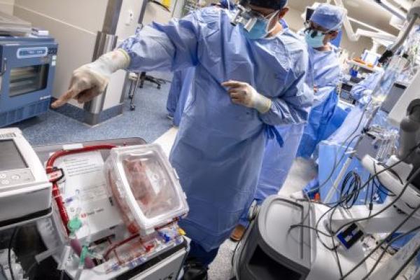 Two surgeons in blue sterile gowns and masks work in a high-tech operating room, standing beside a medical device containing a donor heart preserved in a transparent transport box while monitoring surgical equipment and screens.