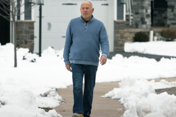 Gentleman walks on a neighborhood sidewalk wearing jeans and a sweater. A house is in the background. Snow covers the yard.