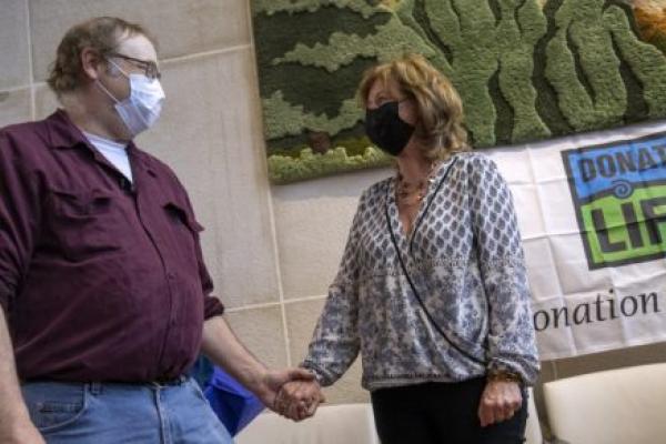 Dwayne Heller, who wears glasses and a face mask and is dressed in a chamois shirt and jeans, clasps hands with Maureen Stathes, who has shoulder length hair and wears a face mask, blouse and pants. A “Donate Life” banner is on the wall behind them.