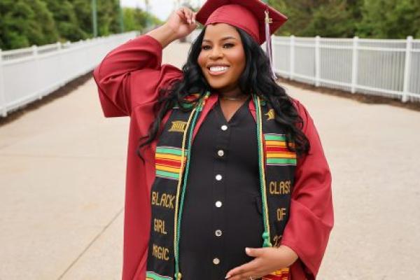 A smiling pregnant woman poses outdoors in a red graduation cap and gown, wearing a black dress and a colorful stole reading “Black Girl Magic” as she cradles her belly.