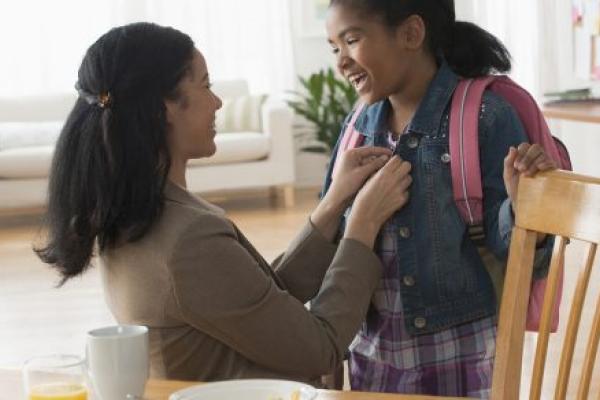 Mother buttons daughter’s jacket as she gets ready for school. Daughter is wearing a backpack. A table with breakfast dishes is in front of them.