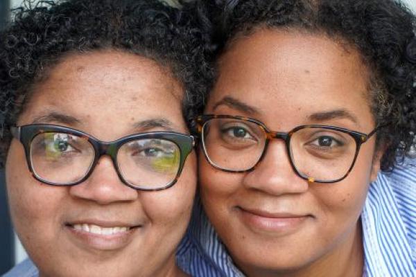 Twin sisters Alana and Alicia Webb smile as they pose in front of a window at their home. Both women have short curly hair and wear glasses and blue shirts.