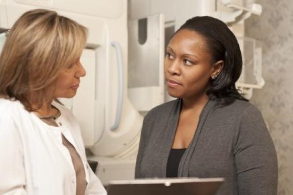 A technician explains a mammogram to an adult woman patient. A mammogram machine is in the background.