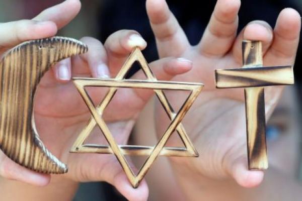 Hands hold large gold symbols associated with religious observances. From left to right, the hands hold the Muslim crescent, the Jewish star, and the Christian cross.
