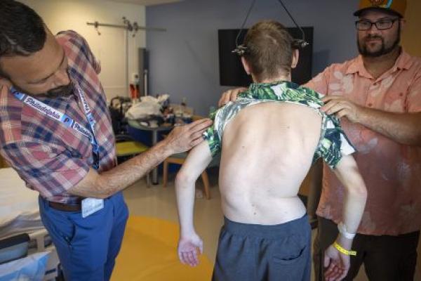 Dr. Brent O'Neill, left, leans over to look at the back of a teen boy with severe scoliosis, while the boy’s father holds the boy’s shirt up. The boy’s spine curves to the left. A halo gravity traction device is attached to his skull. They are standing in a hospital room.