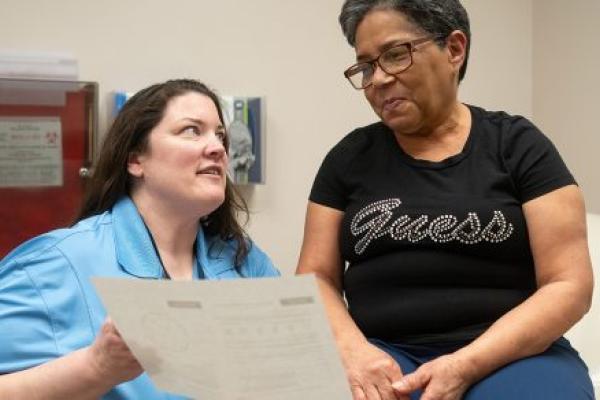 A doctor holds a piece of paper toward a woman patient who is seated on an exam table. The doctor is wearing a blouse with a lanyard around her neck. The woman patient is wearing glasses and a T-shirt with the “Guess” logo on it.