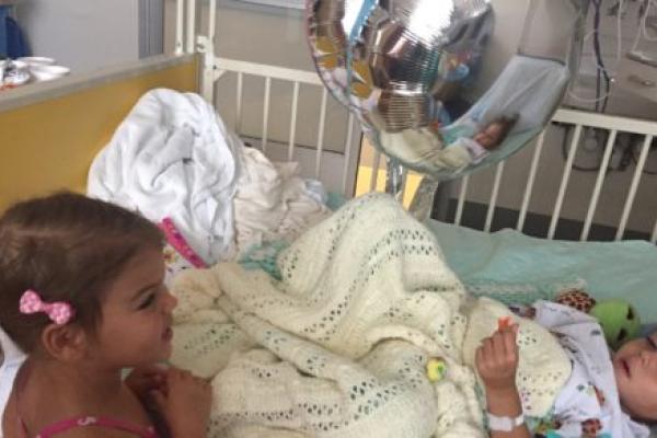 A young girl with pigtails and a ribbon in her hair visits another young girl in a hospital bed.
