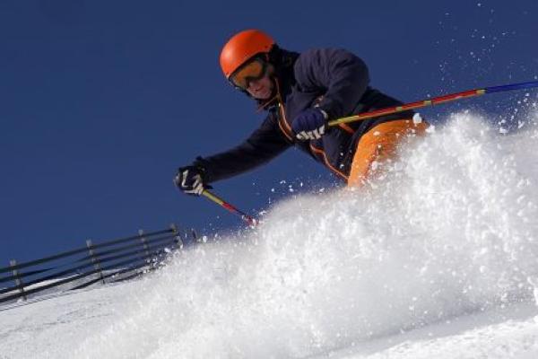 Male skier rides in the deep snow on a sunny day at a ski resort. He is wearing a helmet. A fence is on his left.
