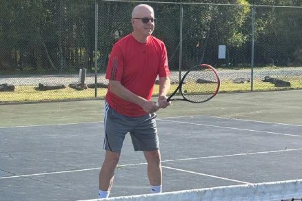Stephen McNamara holds a tennis racket and prepares to return a shot on an outdoor tennis court, with a fence visible in the background.