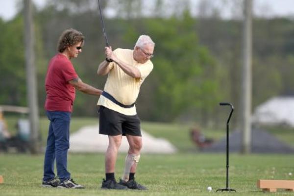 A man swings a golf club while wearing a gait belt and leg brace on his left leg. A man on the left holds his gait belt to give him balance support. A cane is to the right of the golfer.