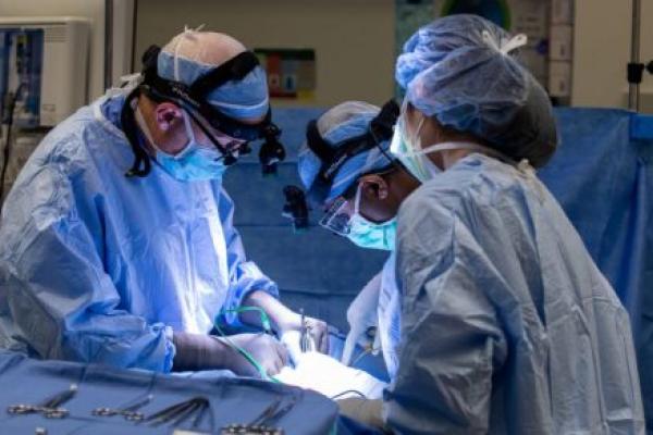 Three medical professionals in surgical gear operate on a patient, not shown. A tray of surgical instruments is within easy reach.