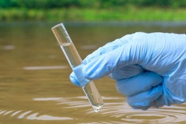 A gloved hand holds a beaker of water above a body of water