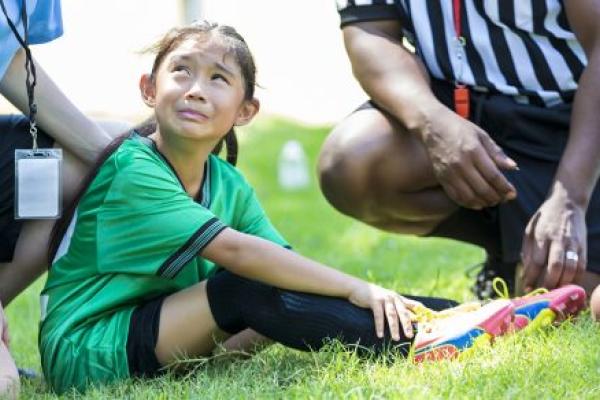 Young female soccer player holds her ankle and grimaces in pain while crying. She is sitting on the playing field with her coach and referee gathered around her.