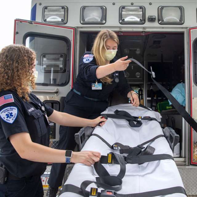 Two female EMTs pull an empty gurney out of the open back doors of an ambulance. Both are wearing navy blue uniforms and face masks. 