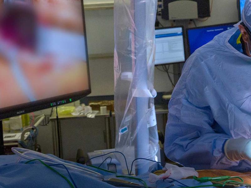 Surgeon in the operating room with full scrubs. He is wearing a cap, mask, glasses and gloves. He is operating on a patient. There is a large light beside him and a screen that shows what he is doing. There are 4 other monitors in the background.
