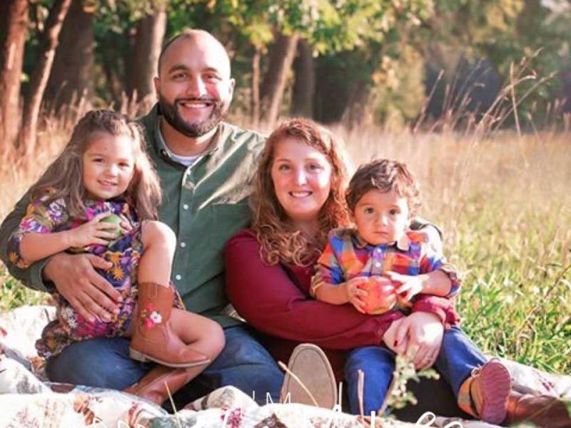 Survivor Aubrey Mora, her husband and two children sitting in a field together on a sunny day.