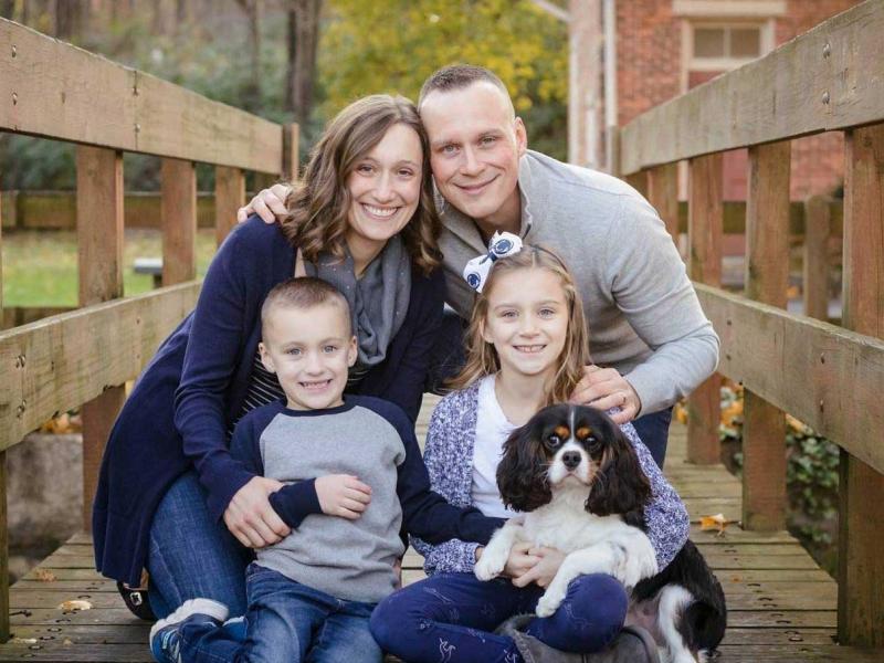 Bradley Heidrich, a 37-year-old survivor of osteogenic sarcoma, is seen with his wife, Julie; daughter, Abby; and son, Ryan. The family of four is pictured standing on a bridge with a creek visible behind them. The two adults are holding the children.