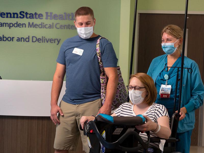 A nurse wearing a COVID mask pushes a masked new mom in a wheel chair. The mom holds a car seat and is smiling at her new baby. The dad, also masked, walks beside them. A registration desk is behind them and a sign above it says Penn State Health Hampden Medical Center Labor and Delivery.