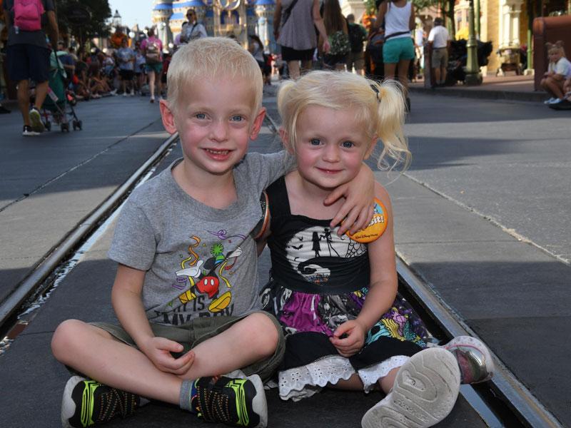 Two small children sit on the ground at Walt Disney World. The boy on the left has his arm around the girl on the right. People can be seen walking and pushing strollers in the background.