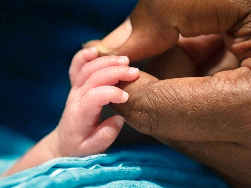 An adult’s hand holds the fingers of an infant as it reaches out from the folds of a blanket.