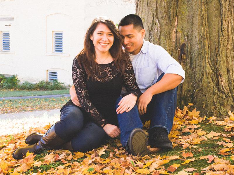 Man and woman smile at camera as they lean in to each other while seated against a tree.
