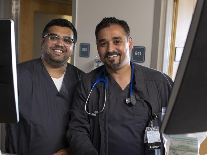 Imran Dawood, left, who is wearing glasses, and his brother, Yasin Dawood, who has a beard, stand side by side behind two medical carts with computer monitors on top. Both men are smiling and are dressed in hospital scrubs. Yasin has a stethoscope around his neck. 
