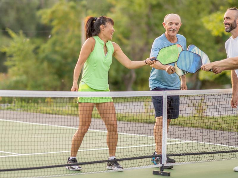 A group of four adults are seen tapping racquets to show sportsmanship after a pickleball game together. They are each dressed comfortably in athletic wear and are smiling as they enjoy their time in the fresh air.