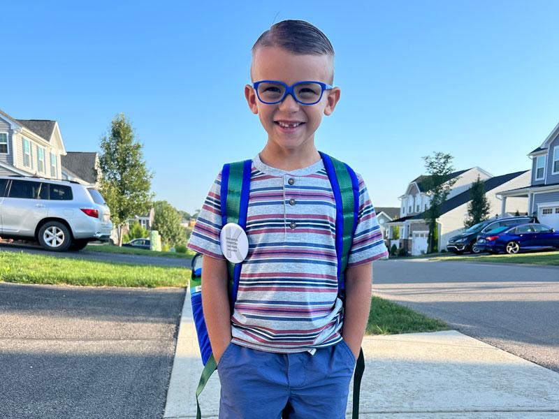Young boy, with hands in his pockets, smiles revealing a missing tooth. He wears glasses and a backpack.