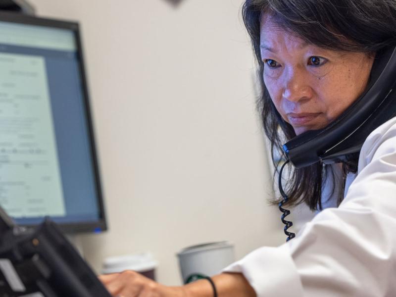 Female medical staff member sitting in front of a computer monitor with a telephone to her ear and reaching with her left to a phone monitor to place a call.