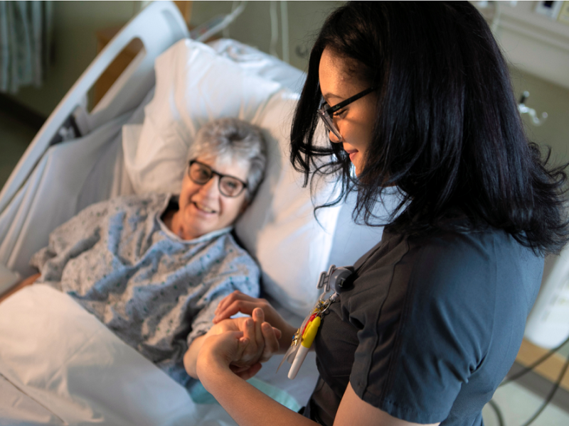 A young female registered nurse with black hair and glasses is standing next to a patient bed. She is holding her patient’s arm and checking her pulse.  The nurse is wearing navy-blue scrubs.