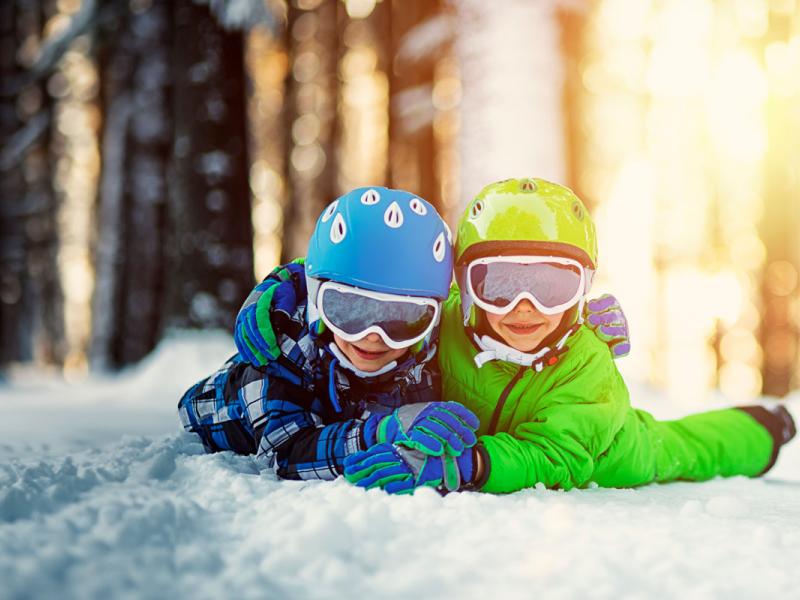 Two children, dressed for cold weather safety, wear snowsuits, gloves, helmets and goggles to keep warm while lying in the snow. In the background the sun filters through the trees.