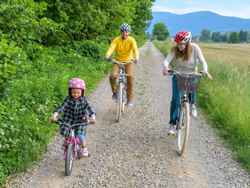 A family models bike safety as they ride together on a country road. Both adults and the child wear a bike helmet.