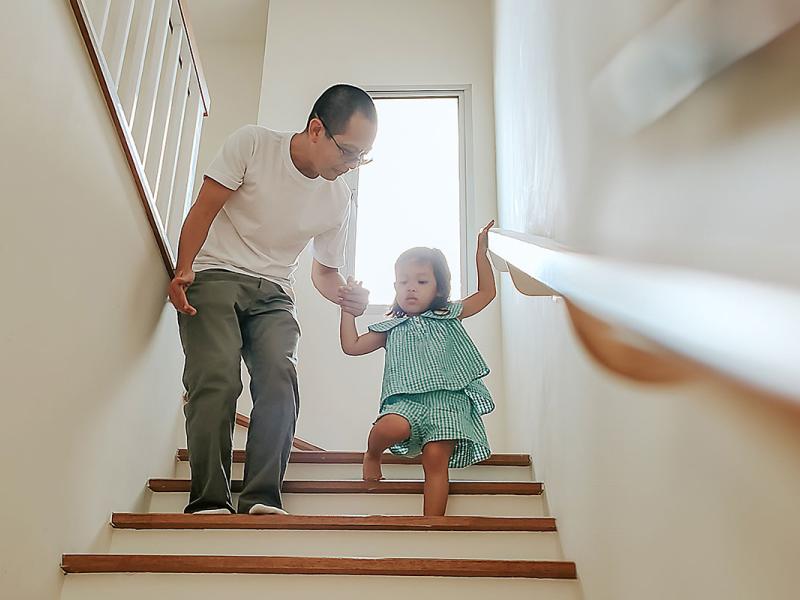 A man is focused on fall prevention as he helps a small child walk down the stairs. The child holds on to a railing with her hand.