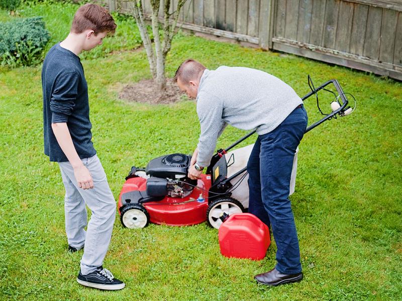 A man grips the pull cord on a lawn mower as he demonstrates lawn mower safety to a teenage boy. They are standing in a yard with bushes, plants and a fence visible in the background.
