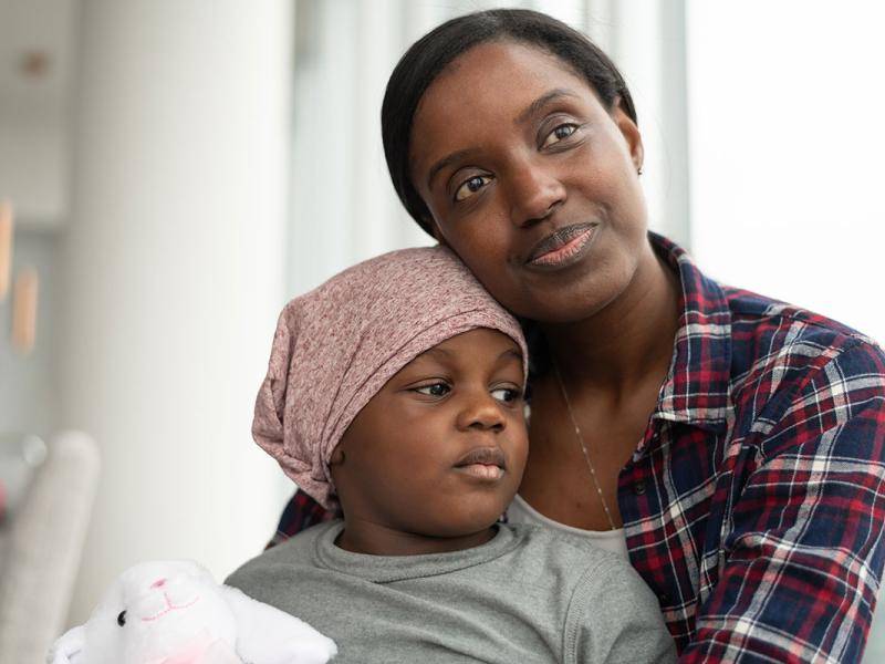 A young black mother lovingly holds her son who is fighting cancer. The child is wearing a bandana and is holding a stuffed rabbit toy. The two individuals are sitting indoors at home near a window. They are filled with gratitude and hope for the future.