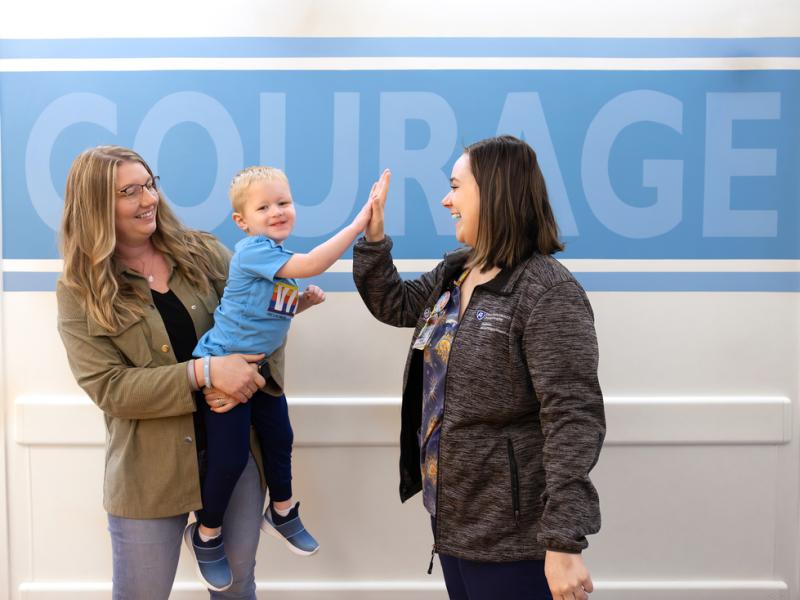 Young child high fiving another adult while their mother holds them standing in front of a sign that says courage.