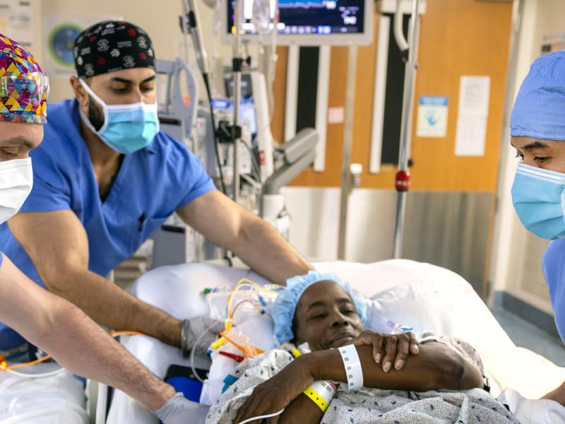 Three registered nurses wearing scrubs and surgical masks move a patient onto an operating room table. Medical monitors, IV lines and hospital equipment are visible in the background.