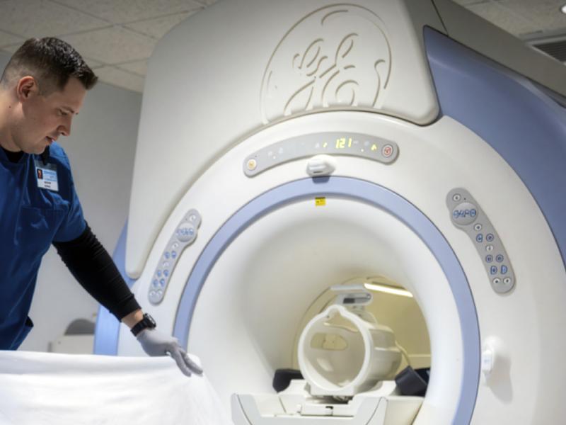 A male imaging tech stands next to an MRI machine and places a sheet onto the table