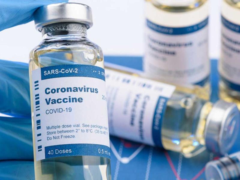 A hand wearing gloves holds a vial of coronavirus vaccine. Three vials are next to the hand. A syringe is in front of them.