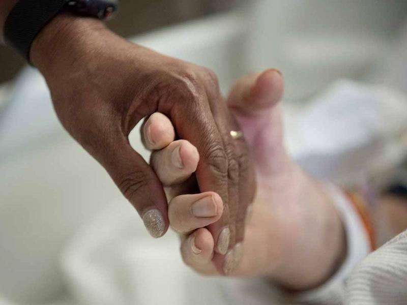 A close-up image shows two hands gently holding each other, with one hand resting on a hospital patient’s hand surrounded by blankets and medical equipment.